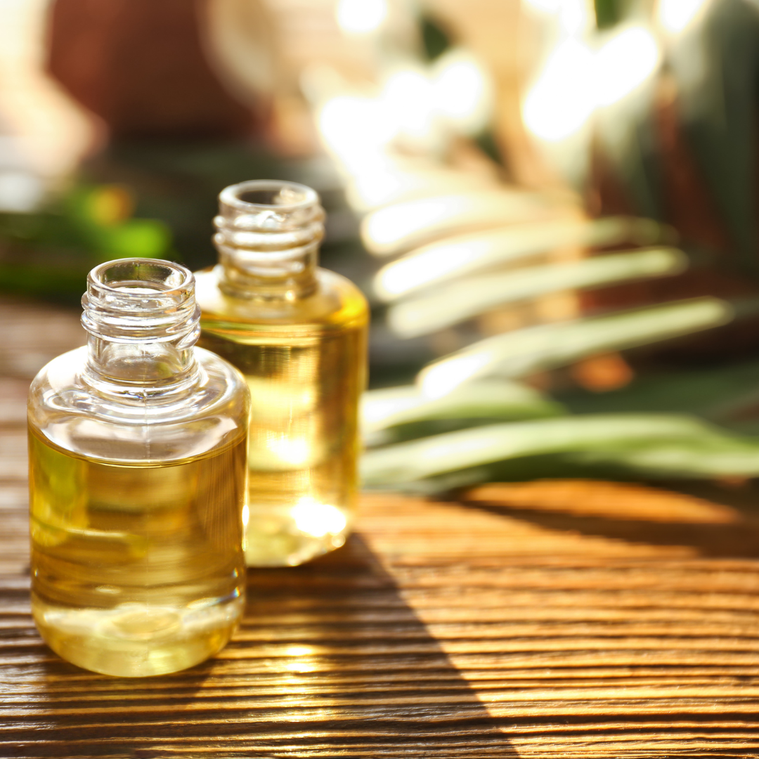 Zwei kleine Glasfläschchen mit goldenem Duftöl auf einer hölzernen Oberfläche, im Hintergrund unscharfe grüne Pflanzen im Sonnenlicht.